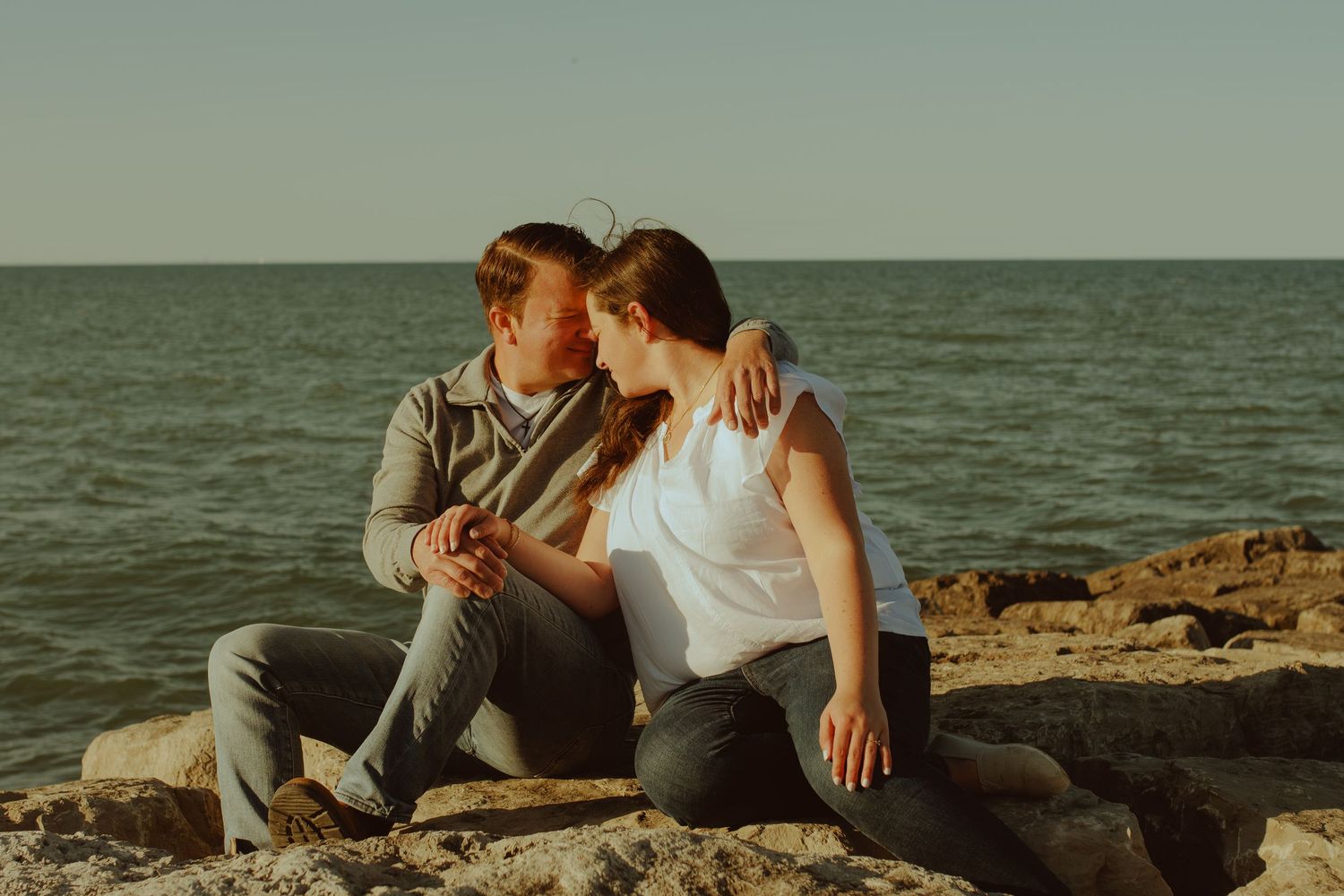 Romantic beach engagement photo session with couple sitting on coastal rocks at sunset near the ocean.