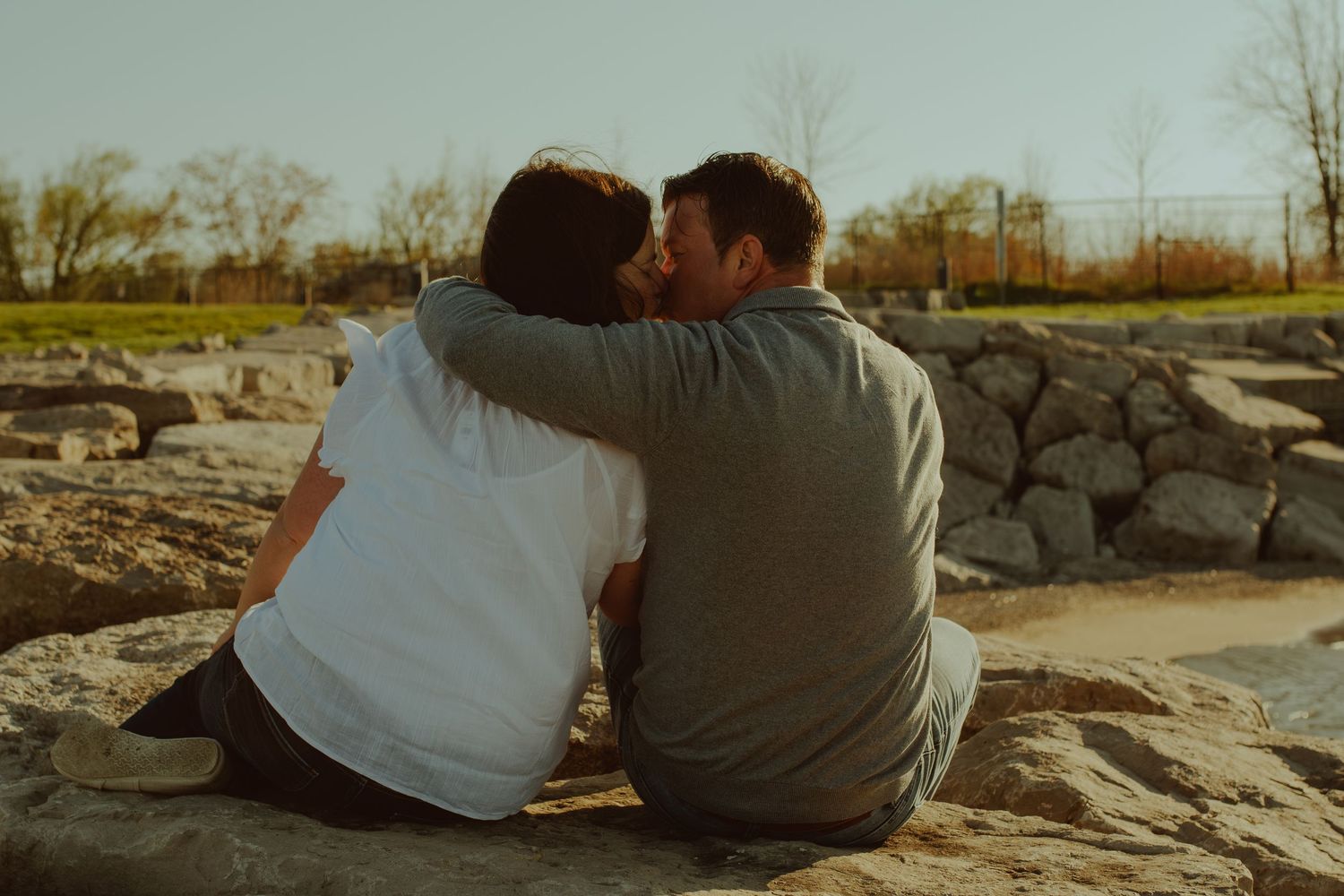Two people sit close together on rocky shore during sunset creating a romantic silhouette against the scenic backdrop.