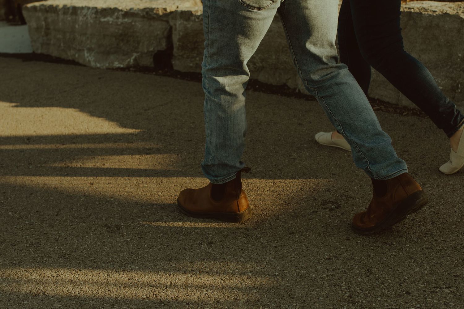 Feet of multiple people walking on a concrete sidewalk in casual shoes at sunset.