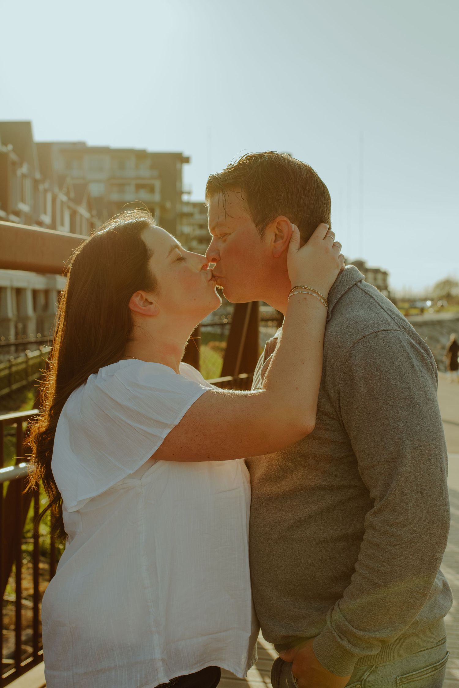 A romantic couple shares tender kisses while embracing in warm sunlight against an urban backdrop.