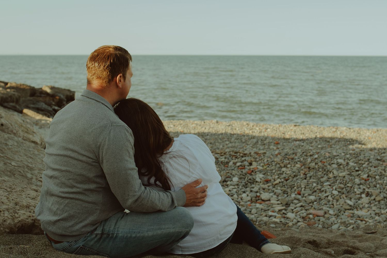 A couple sits together on a pebble beach overlooking the ocean at sunset.
