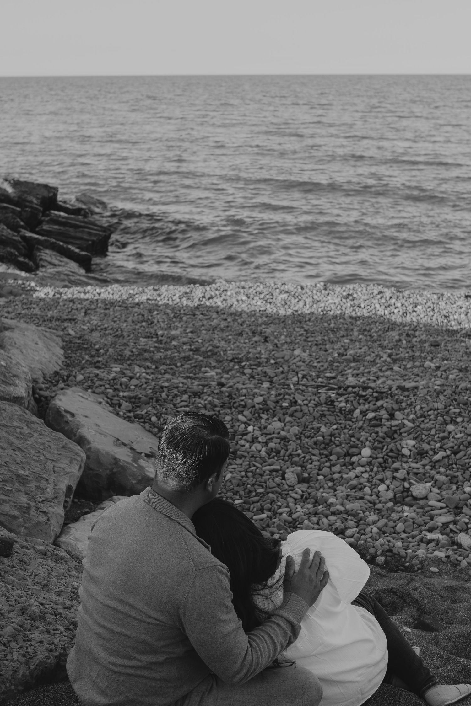A couple sits together on rocky coastline overlooking dark ocean waves in black and white.