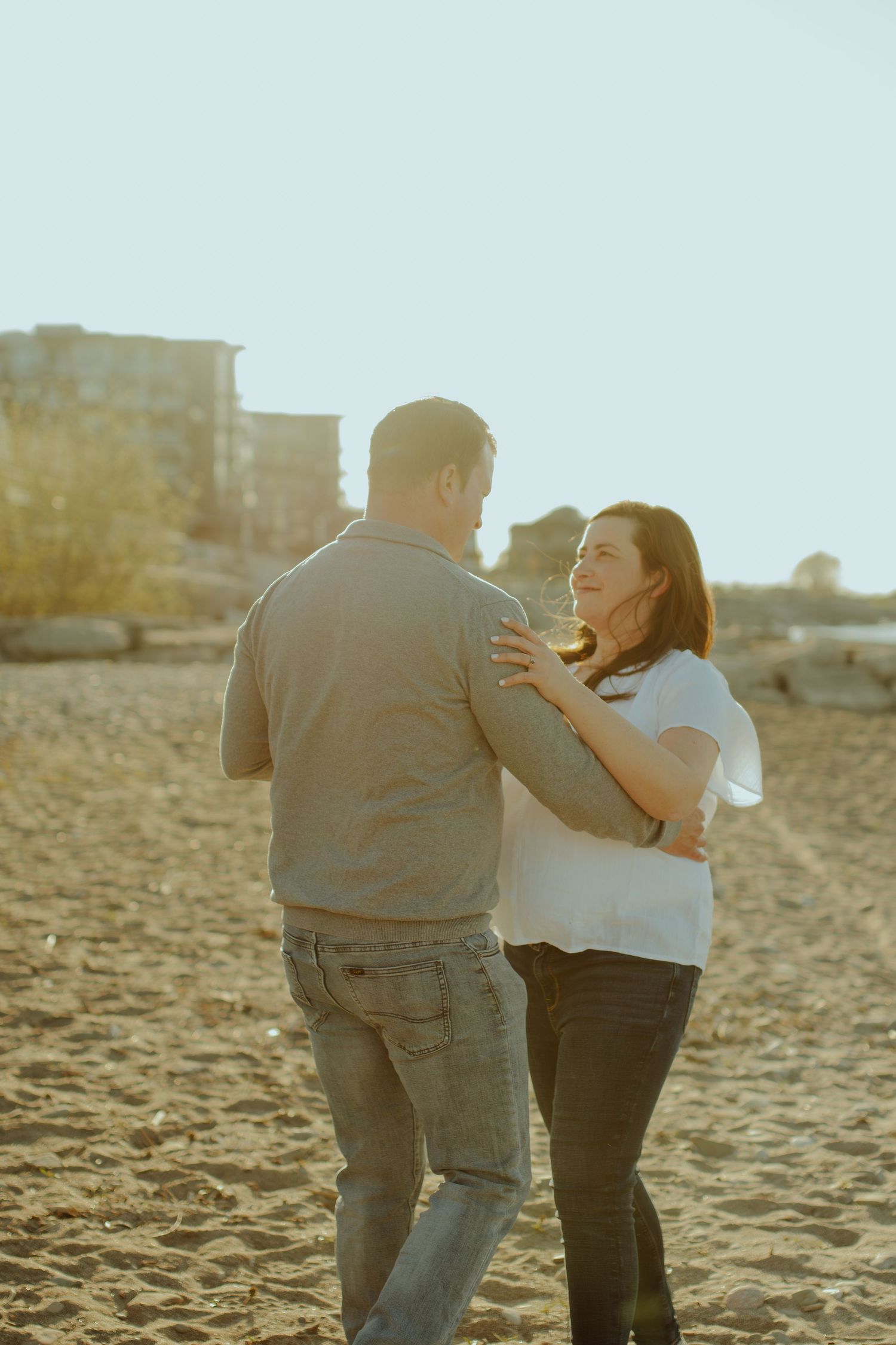 A couple walks along a sandy beach together during golden hour with buildings in the background.