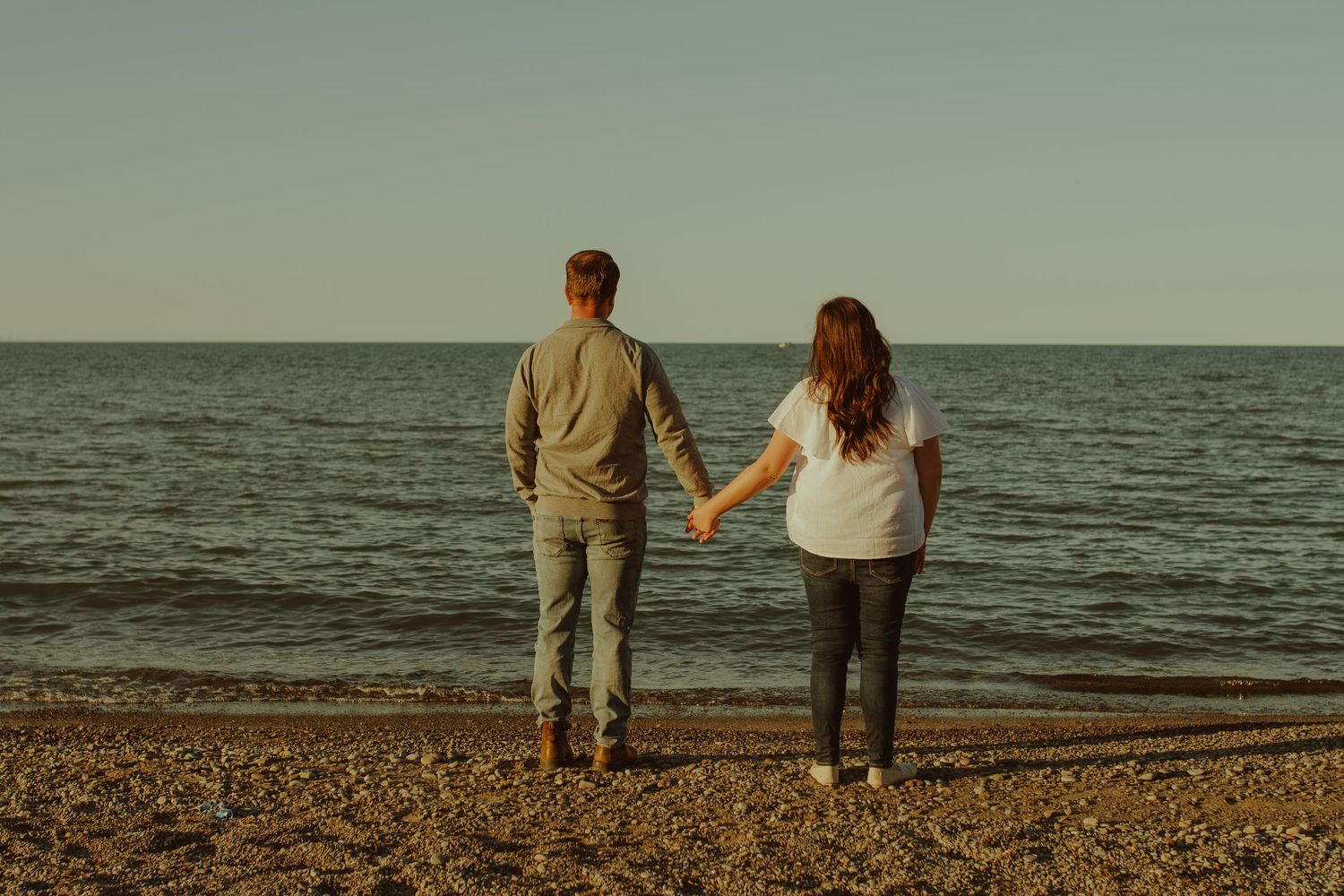 Couple standing together on pebble beach overlooking ocean at sunset.
