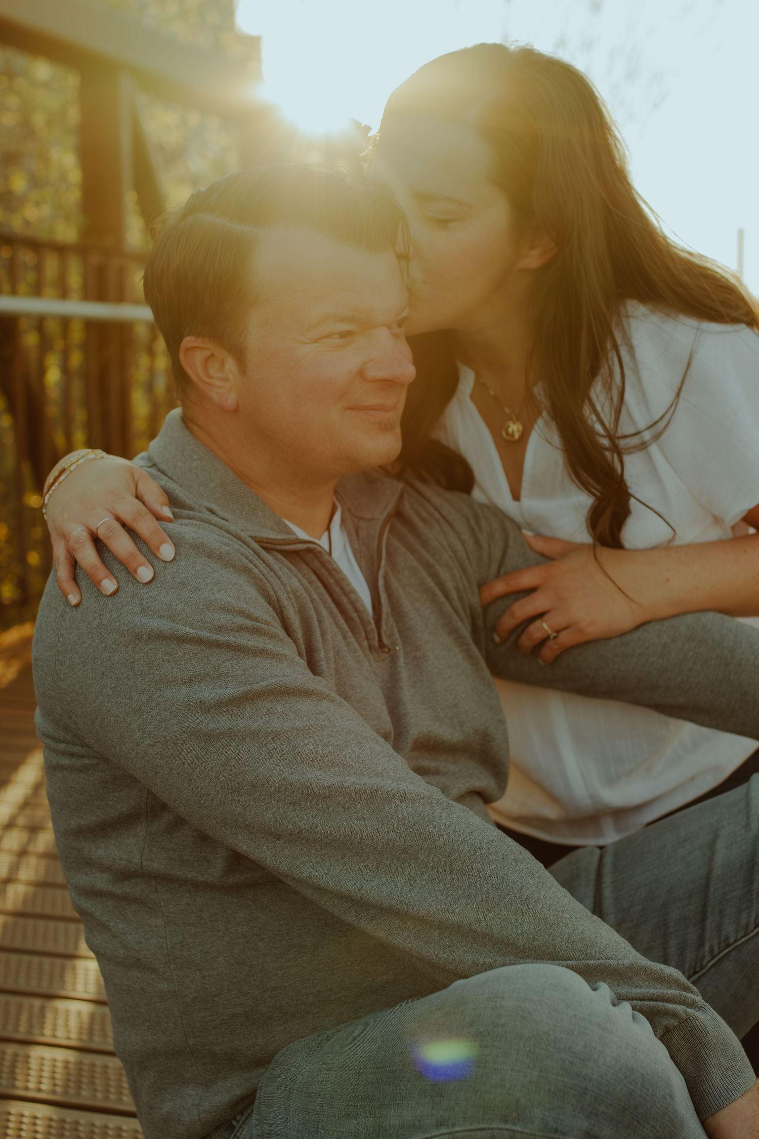 A couple sits close together on outdoor steps during golden hour, sharing an intimate moment in warm sunlight.