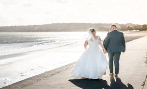 A couple walks along a scenic beach shoreline at sunset in elegant wedding attire.