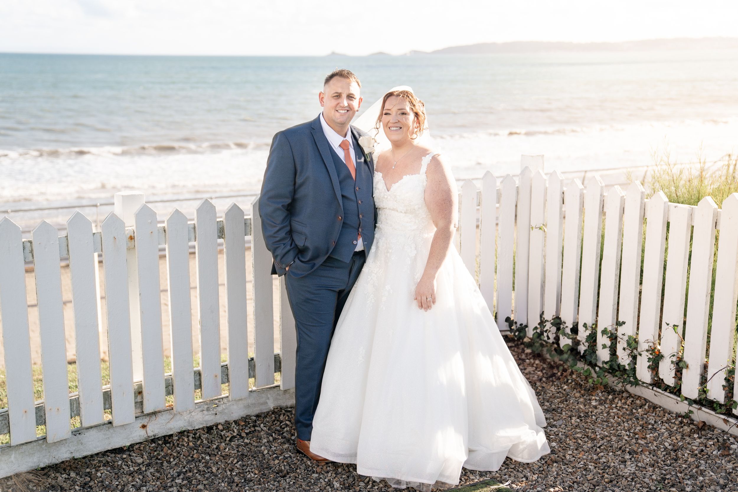 Newlyweds share an intimate moment while walking along a scenic beachfront with a white picket fence.