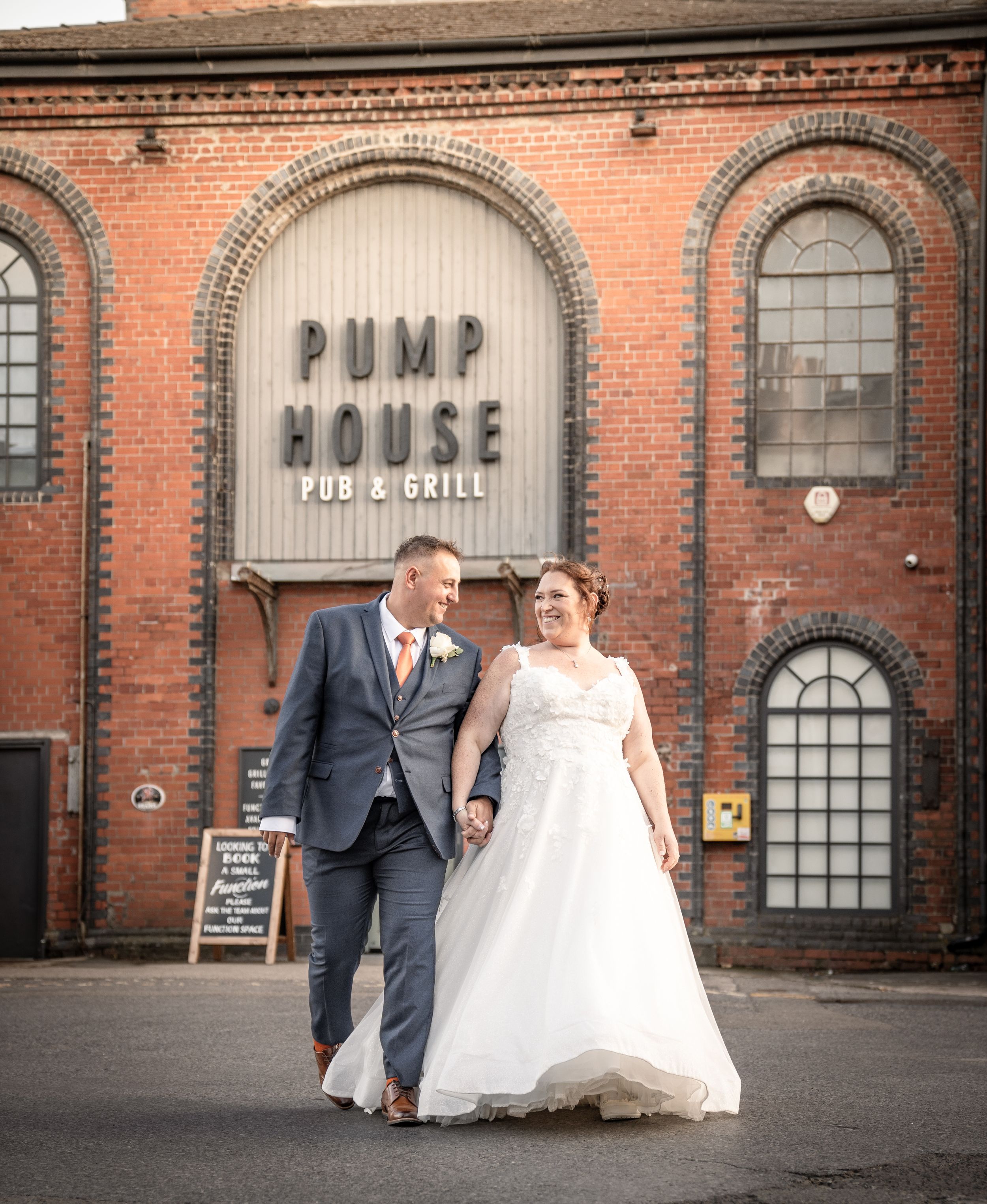 A couple walks together in front of a historic red brick building with arched windows and industrial architecture.