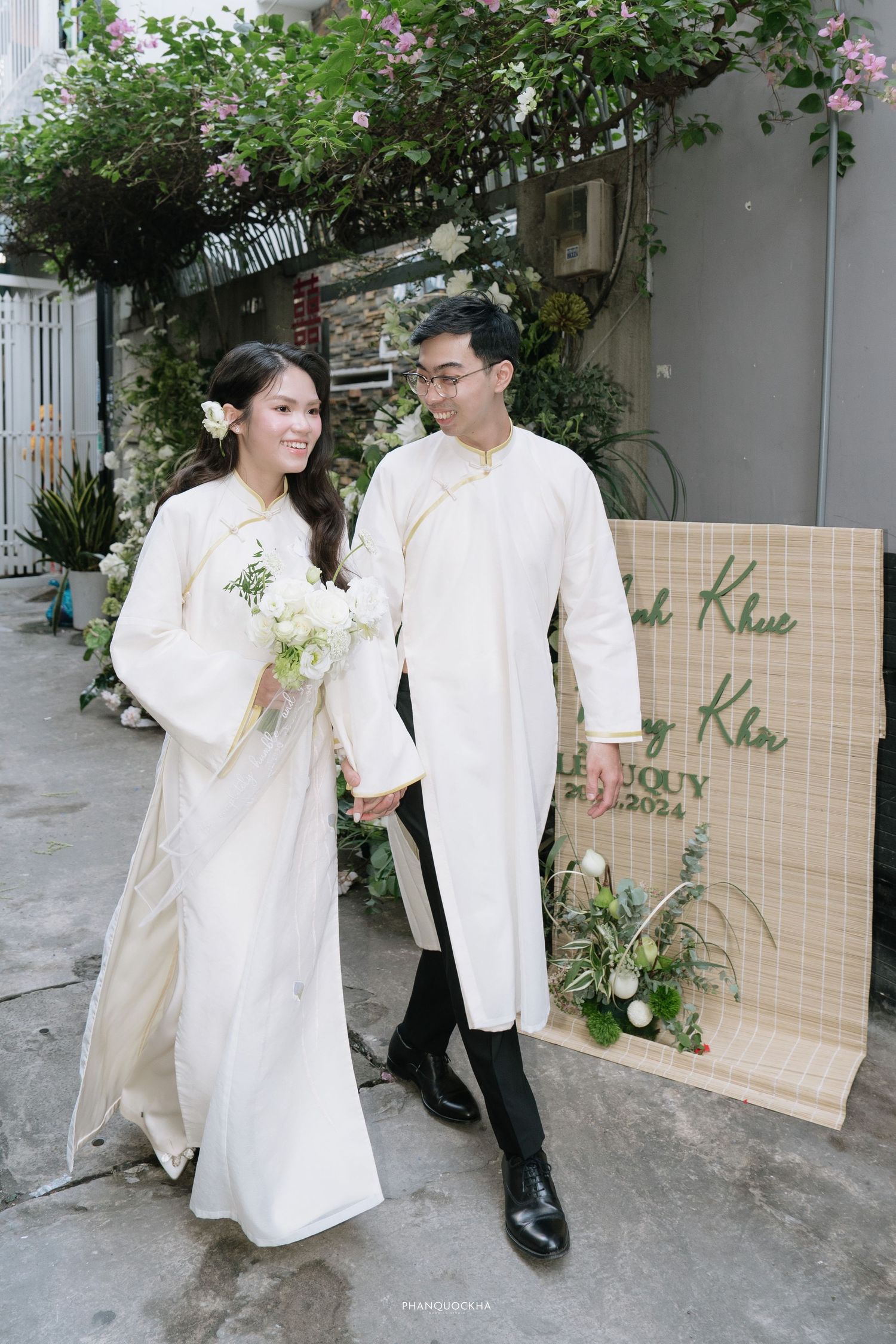 Smiling Vietnamese bride and groom in white ao dai hold hands at outdoor wedding ceremony.