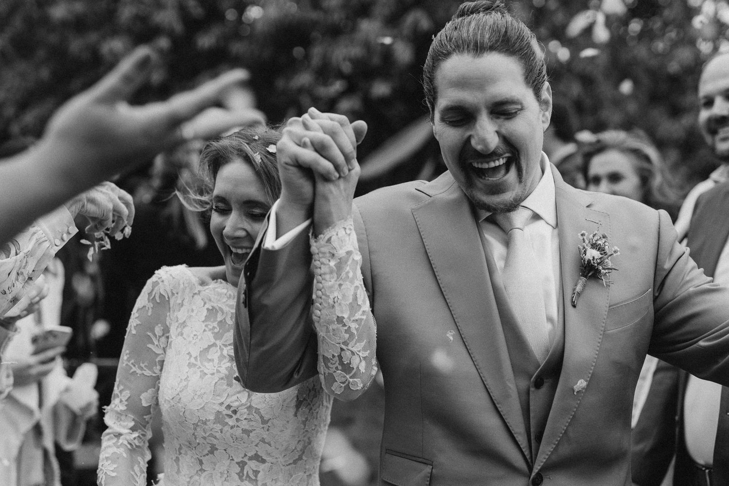 A joyful bride and groom laugh together surrounded by happy guests during an outdoor wedding celebration.