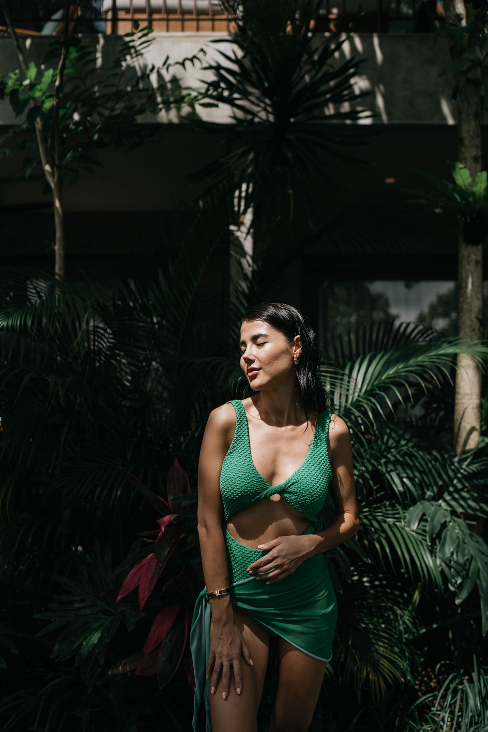 Woman in green swimwear poses against tropical foliage in moody lighting.