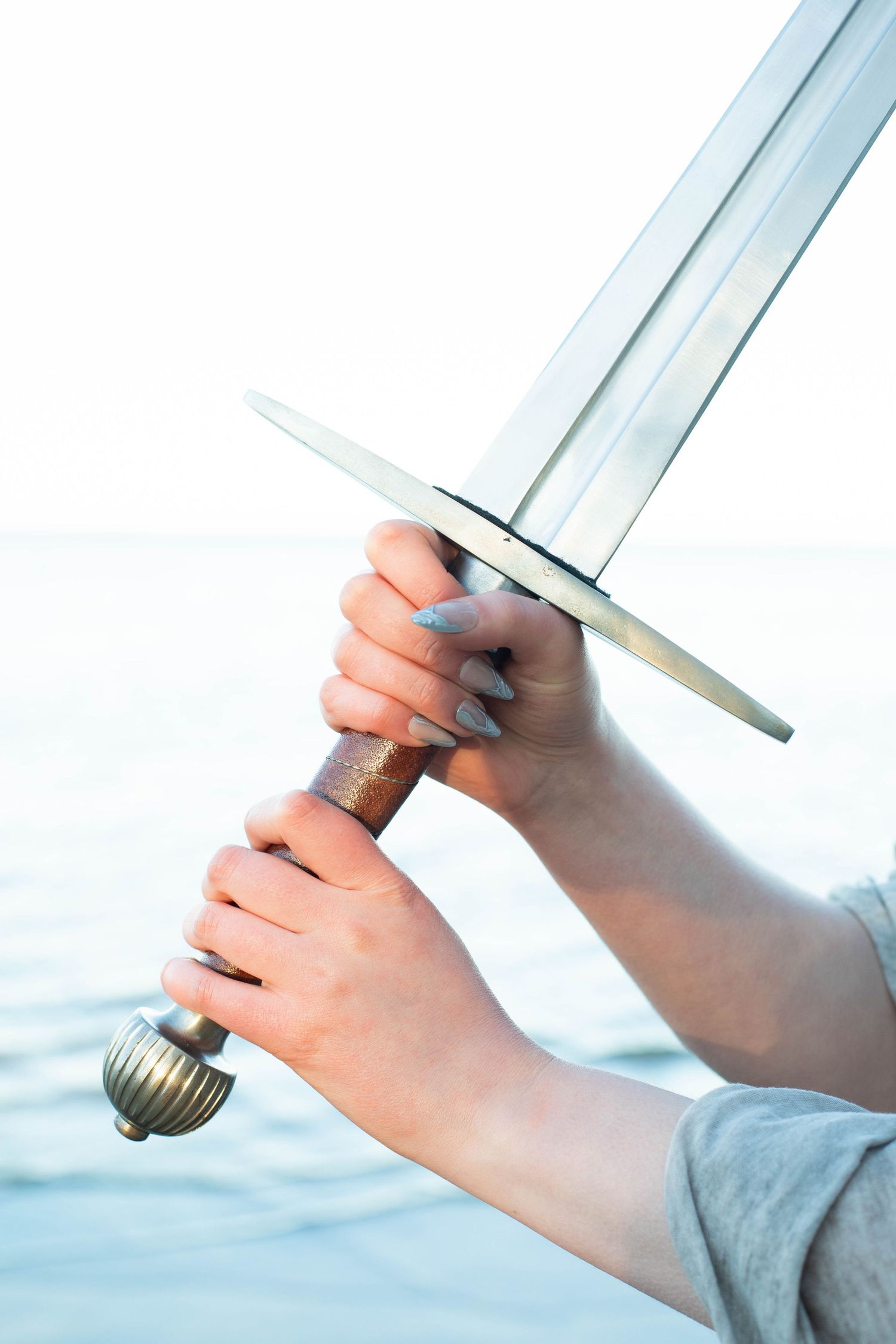 Person with cute nails holding a sword by a body of water.