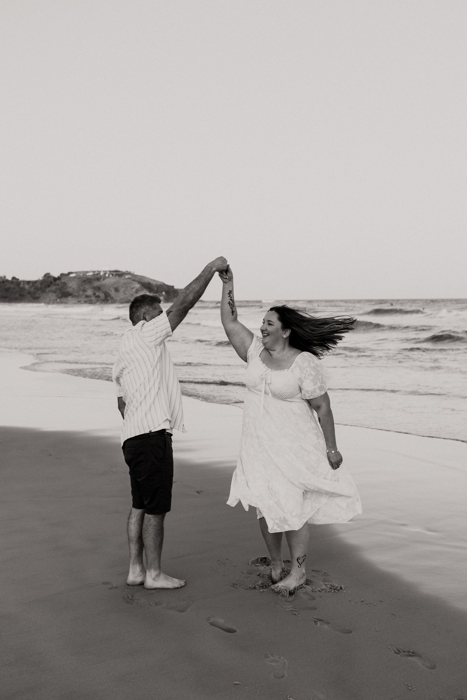 A couple in black and white dances playfully together on a beach with ocean waves in the background.