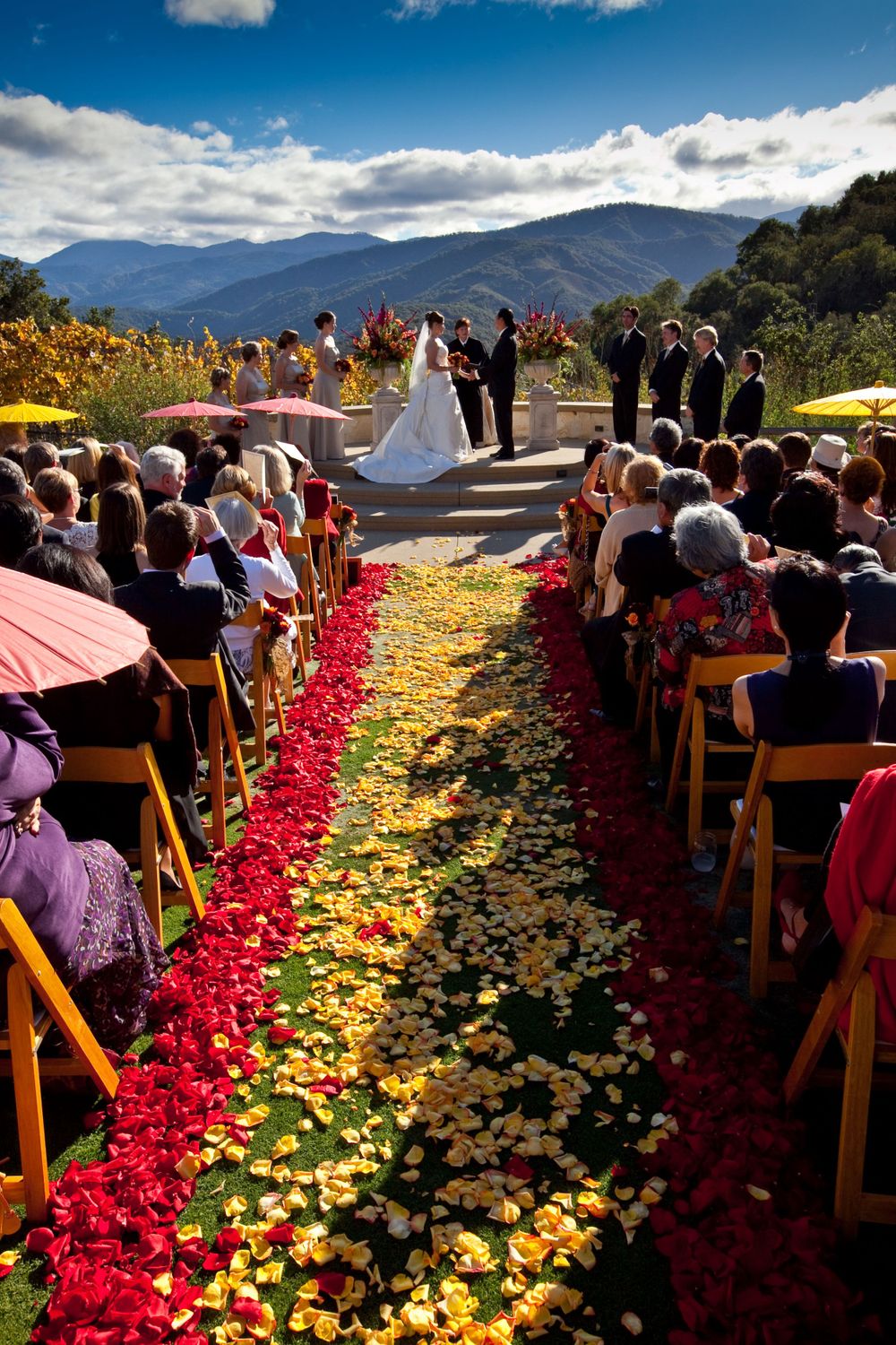 Outdoor mountain wedding ceremony with red and yellow rose petals lining the aisle against a dramatic mountain backdrop.