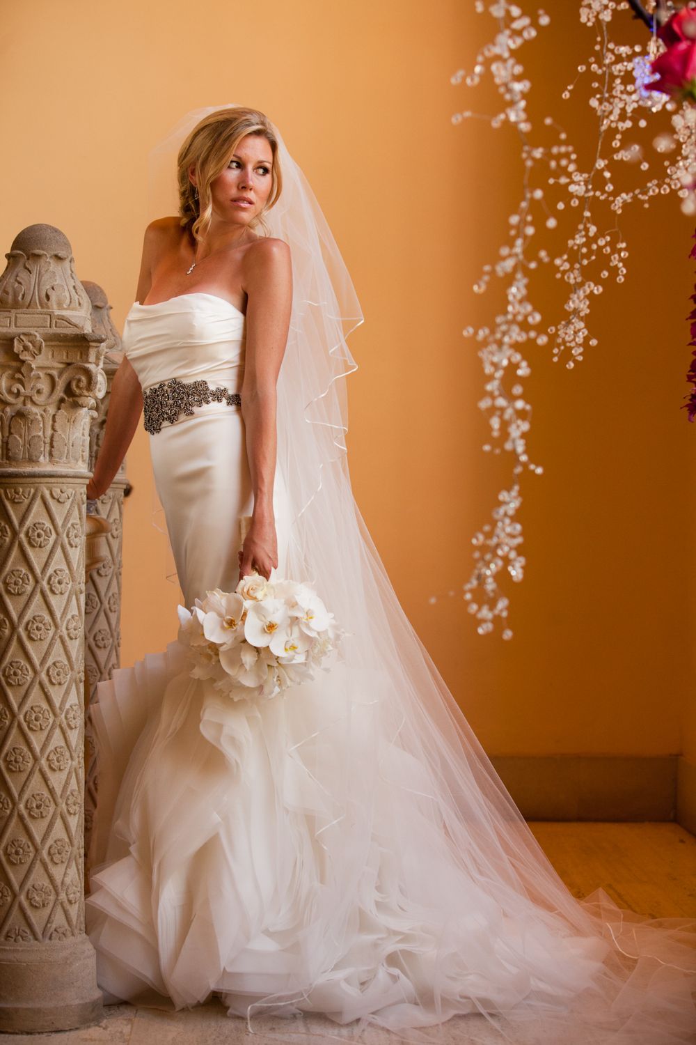 An elegant wedding dress with crystal details and flowing train beside an ornate stone column.