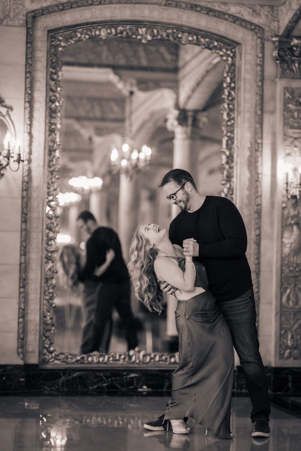 A couple dances gracefully in an elegant ballroom with ornate mirrors and chandeliers.