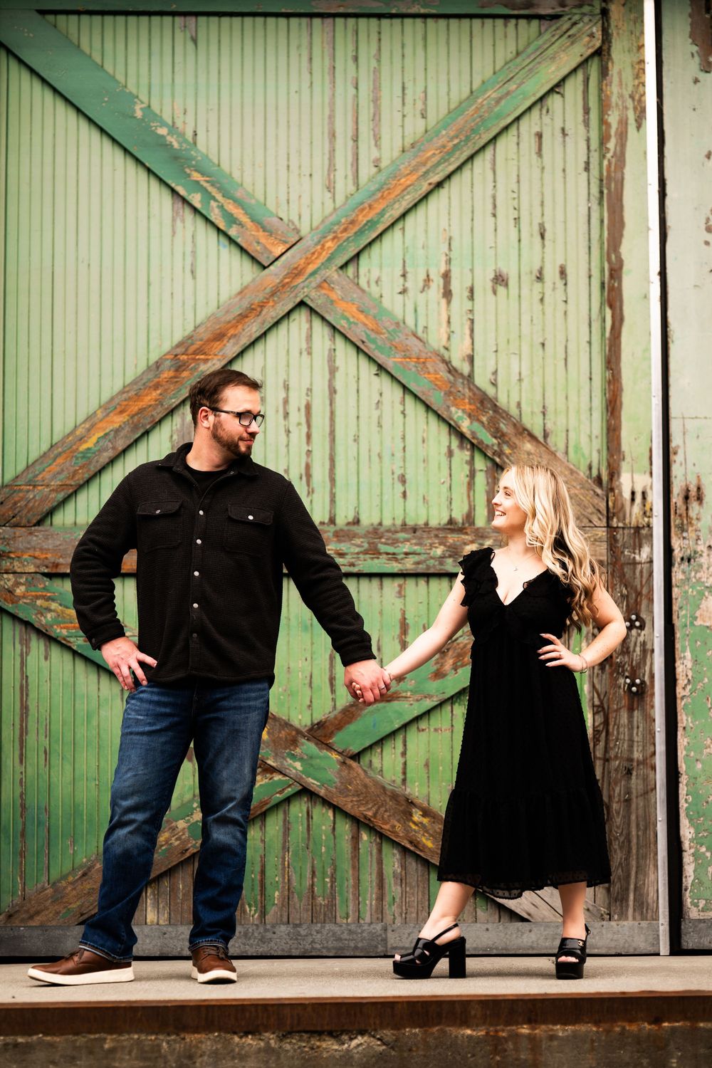 A stylish couple poses against weathered green barn doors in a rustic setting.