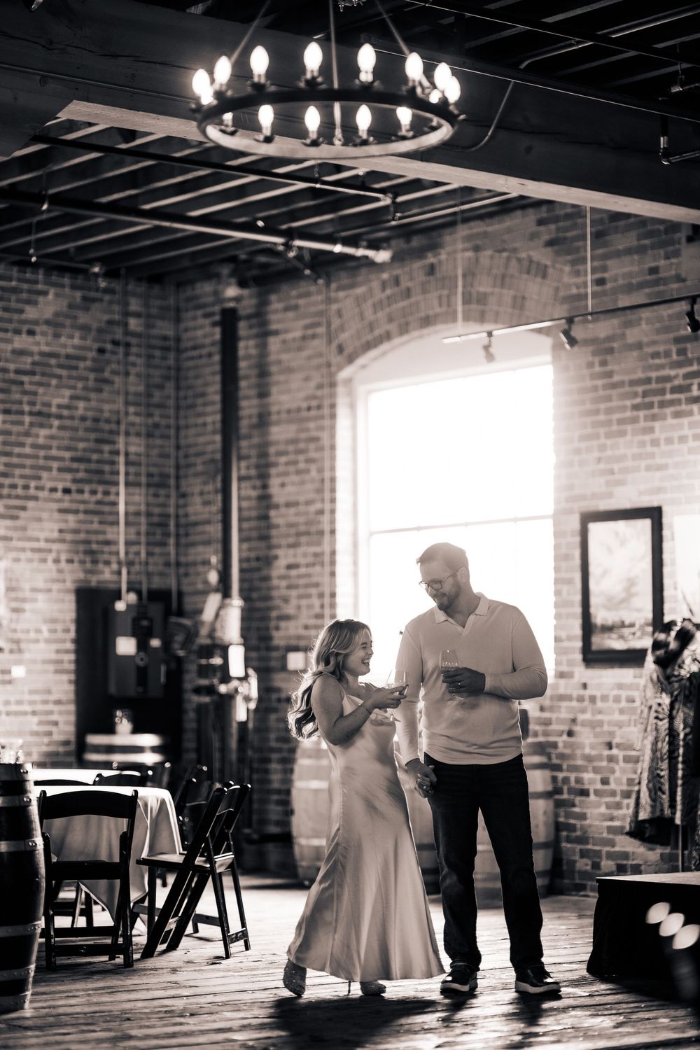 A romantic couple dancing together in an industrial brick-walled venue with elegant chandeliers overhead.