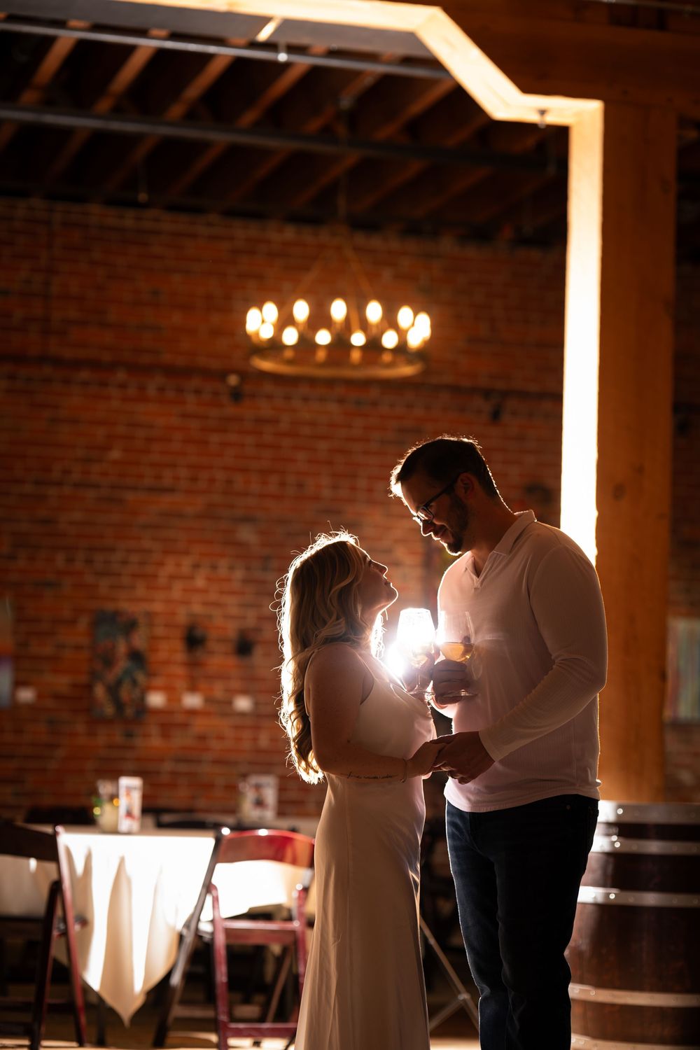 A silhouetted couple shares an intimate moment in a rustic venue with exposed brick walls and ambient chandelier lighting.