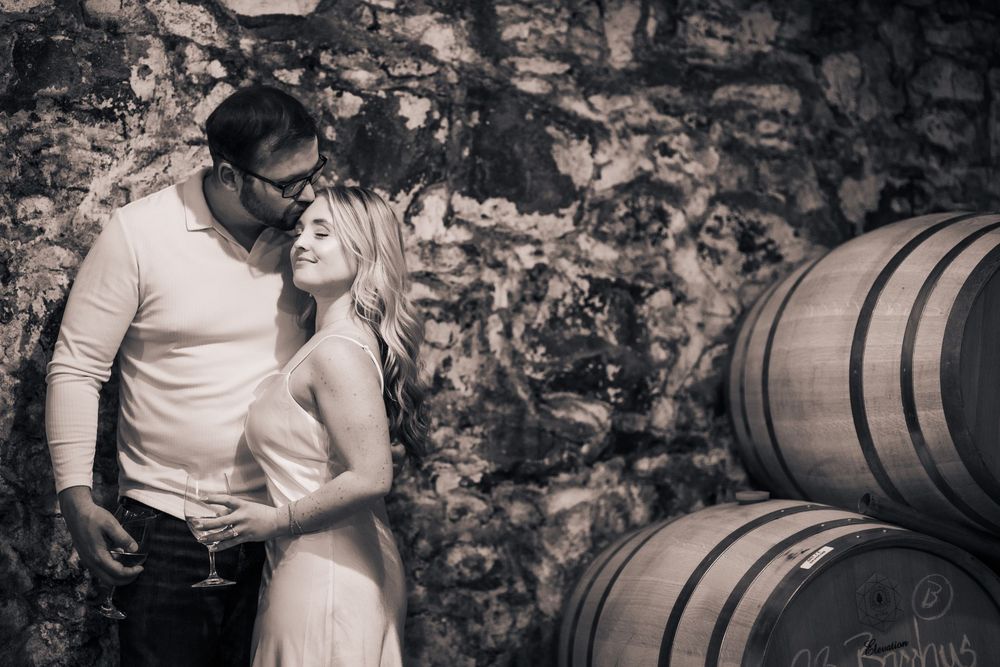 A romantic black and white photo of a couple embracing next to wine barrels in a rustic stone cellar.