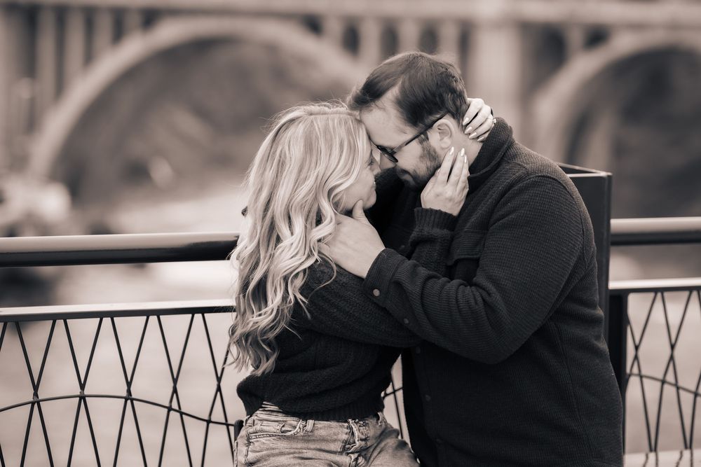 A romantic couple shares an intimate moment on a bridge railing in a black and white toned photograph.