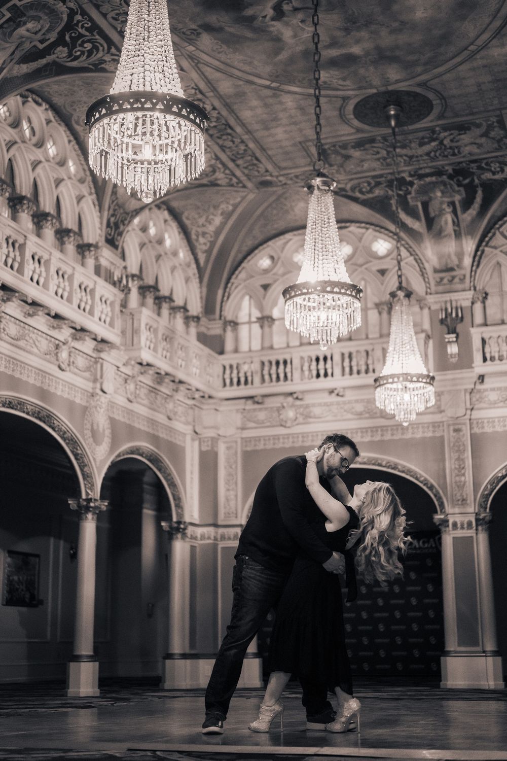 A romantic couple dances in an elegant ballroom with ornate architecture and crystal chandeliers overhead.