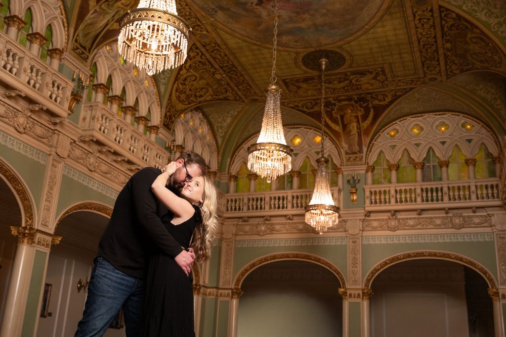 A couple shares an intimate dance moment beneath sparkling chandeliers in a grand historic ballroom.