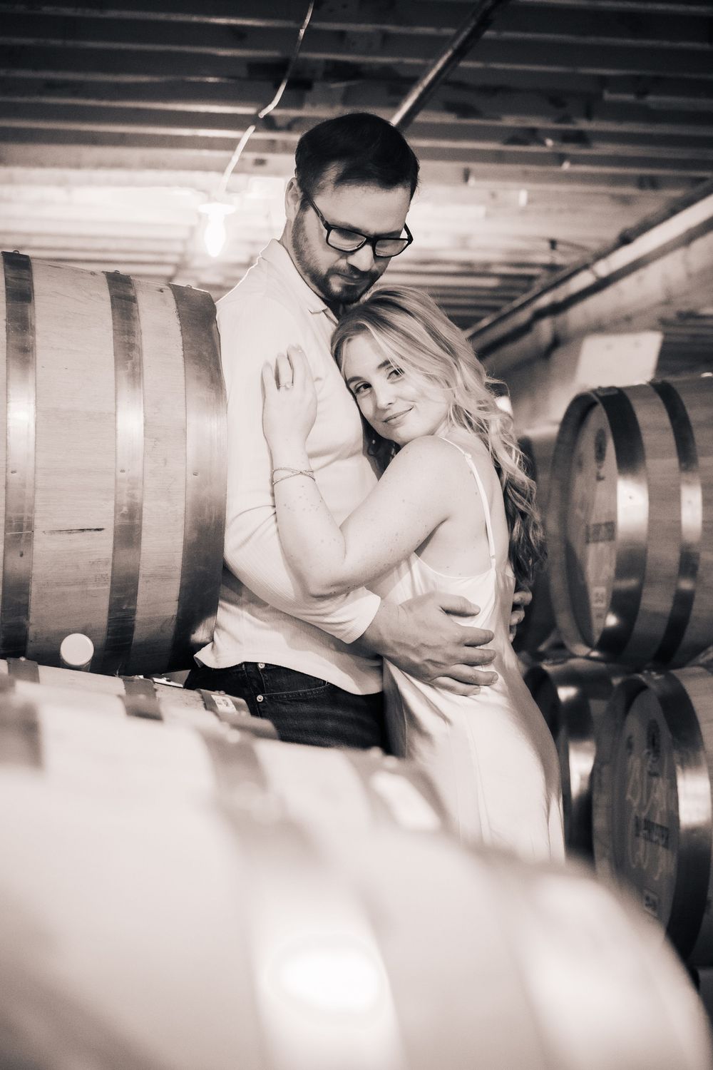 Couple embracing in a rustic wine cellar with wooden barrels in the background.