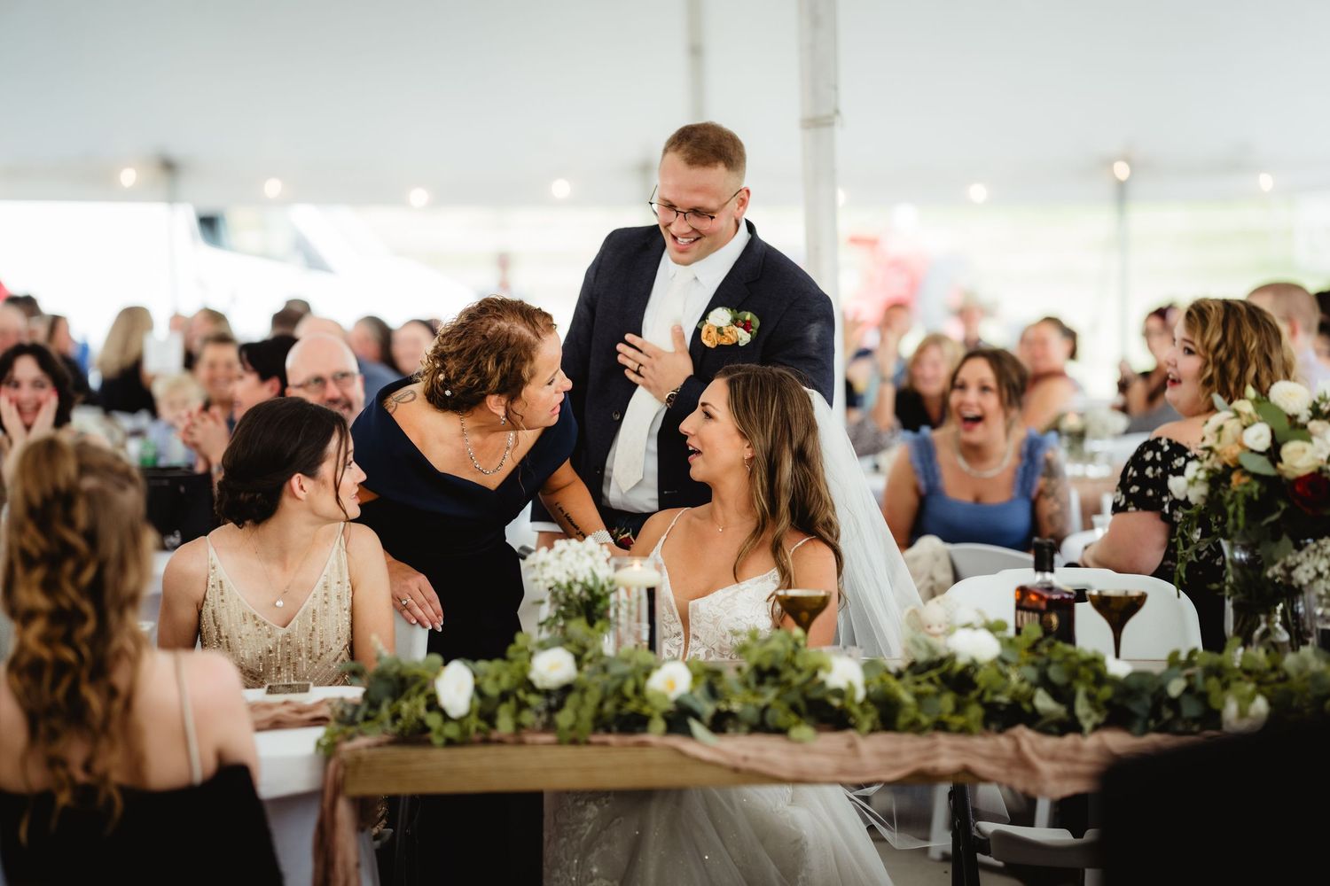Wedding reception guests gathered at an elegantly decorated table with floral centerpieces inside a white tent pavilion.