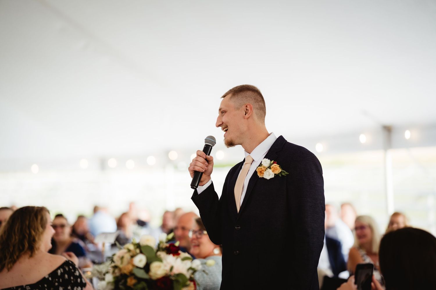 A groom gives an emotional speech at an indoor wedding reception with floral arrangements and guests in the background.