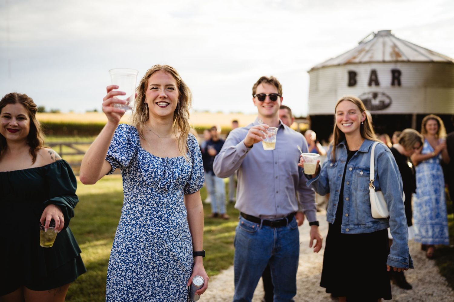 Friends cheering with drinks at an outdoor gathering near a rustic barn and grain silo at sunset.