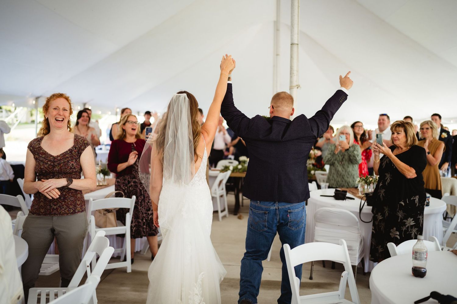 Wedding guests dance and celebrate with raised arms under a white tent during reception festivities.