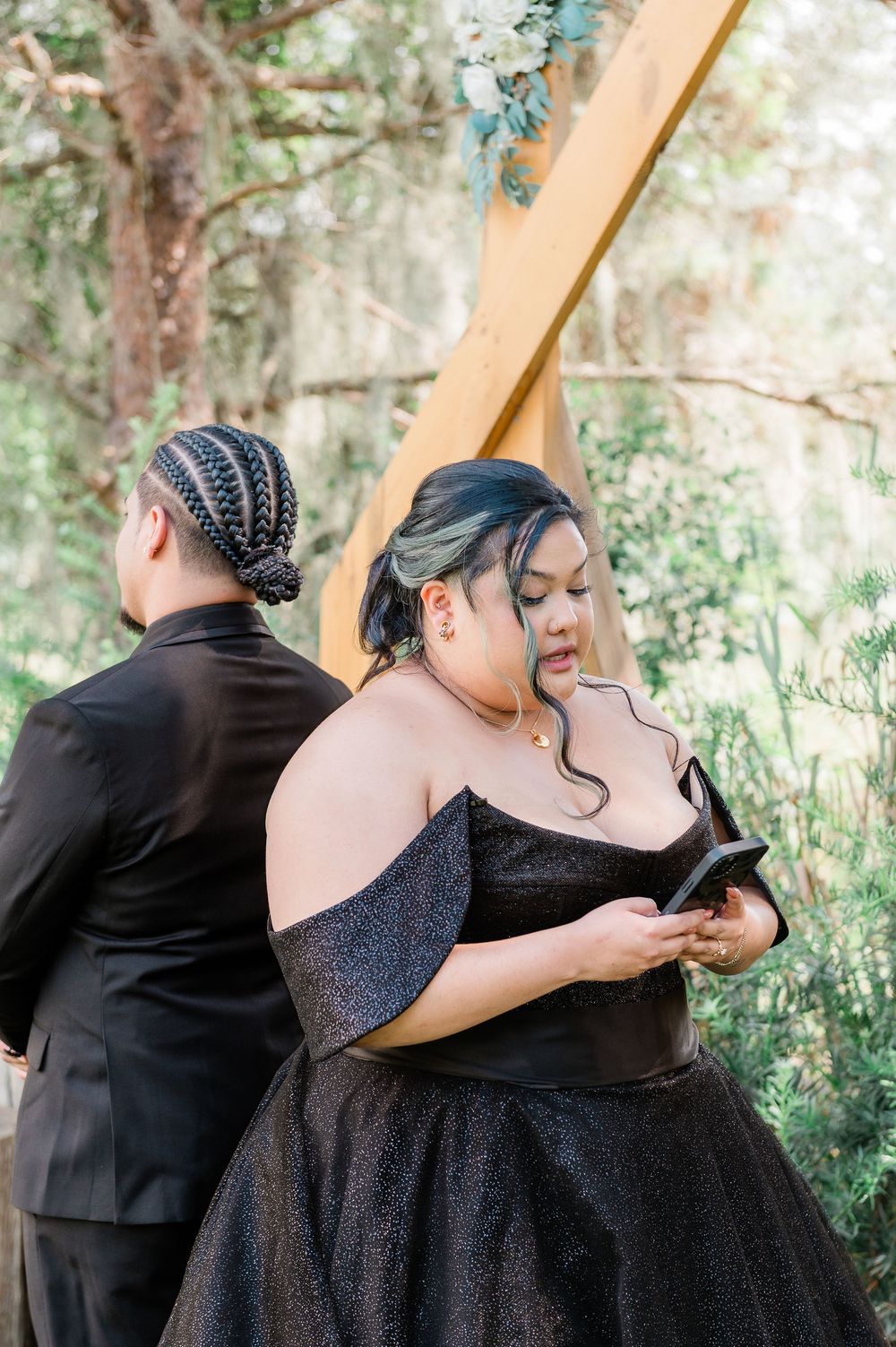 A couple in formal black attire embracing in a romantic photoshoot series among trees and natural scenery.