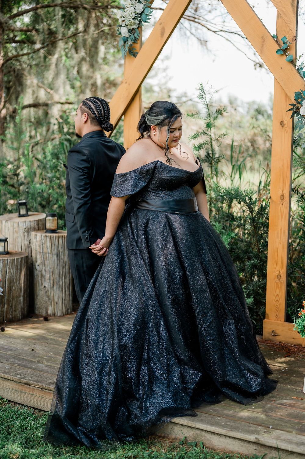 Black lace ballgowns flow elegantly against wooden pillars in a garden setting during a formal photo session.