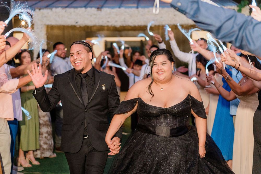 A couple in black formal attire exits through a sparkler send-off at their wedding celebration.