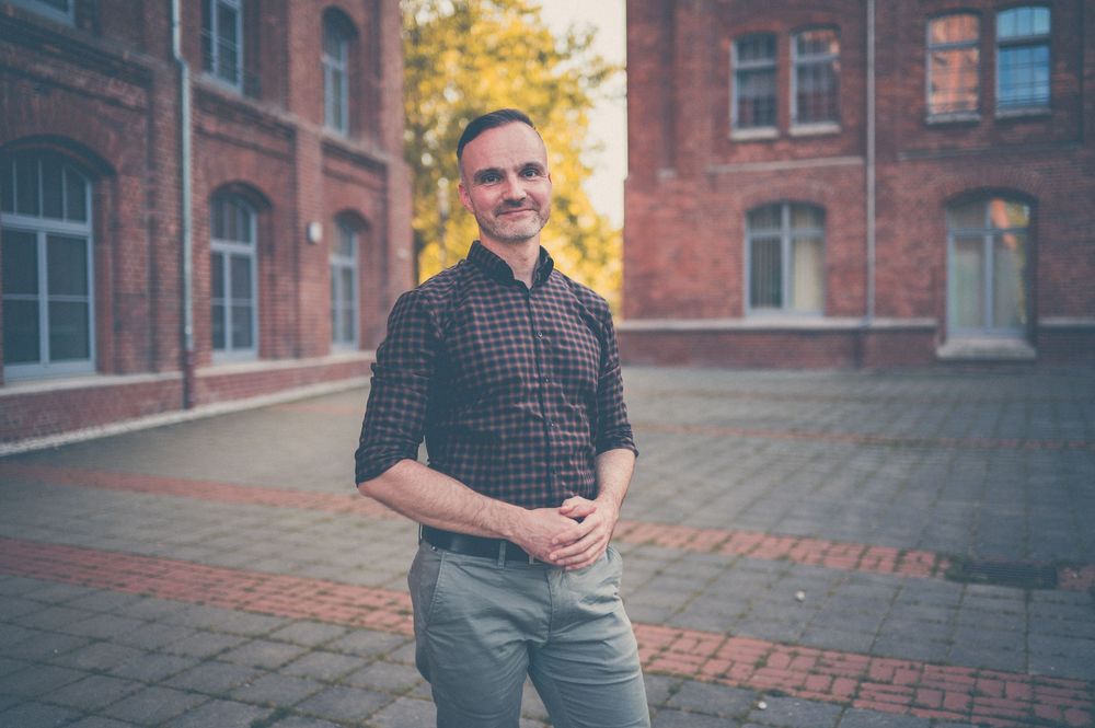 A person in a plaid shirt poses outdoors near red brick buildings during autumn with golden foliage visible.
