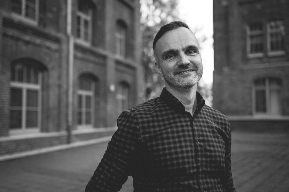 Professional headshot photo in front of brick building with checkered jacket and warm smile.