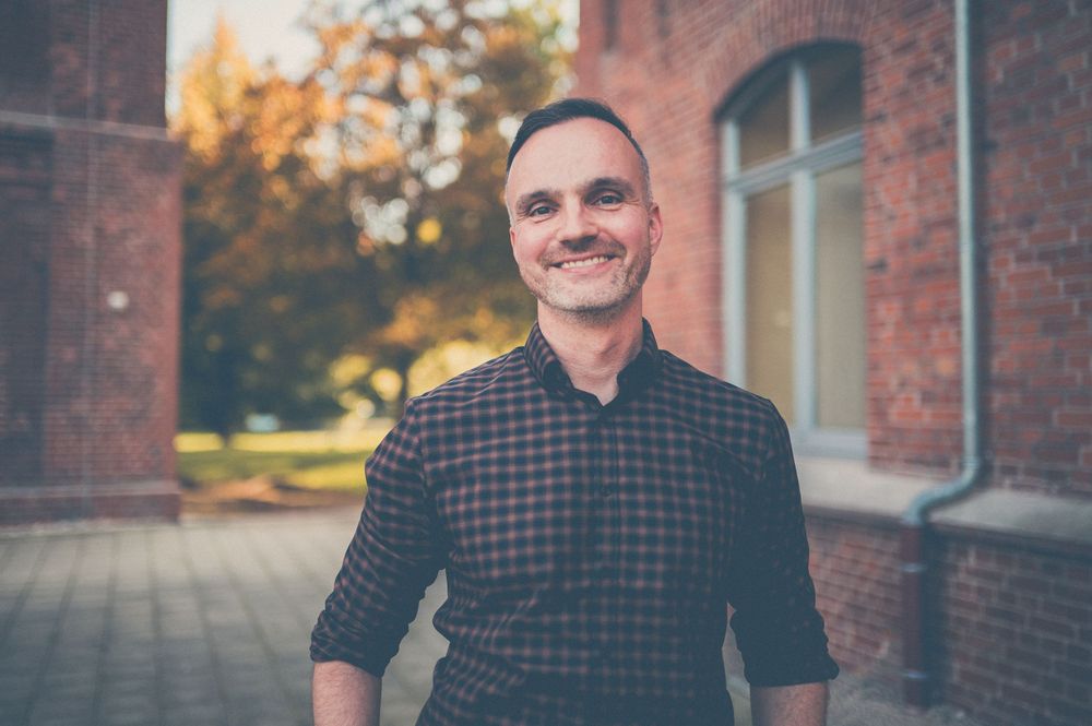 Someone in a checkered button-down shirt smiles warmly outside a brick building on an autumn day.