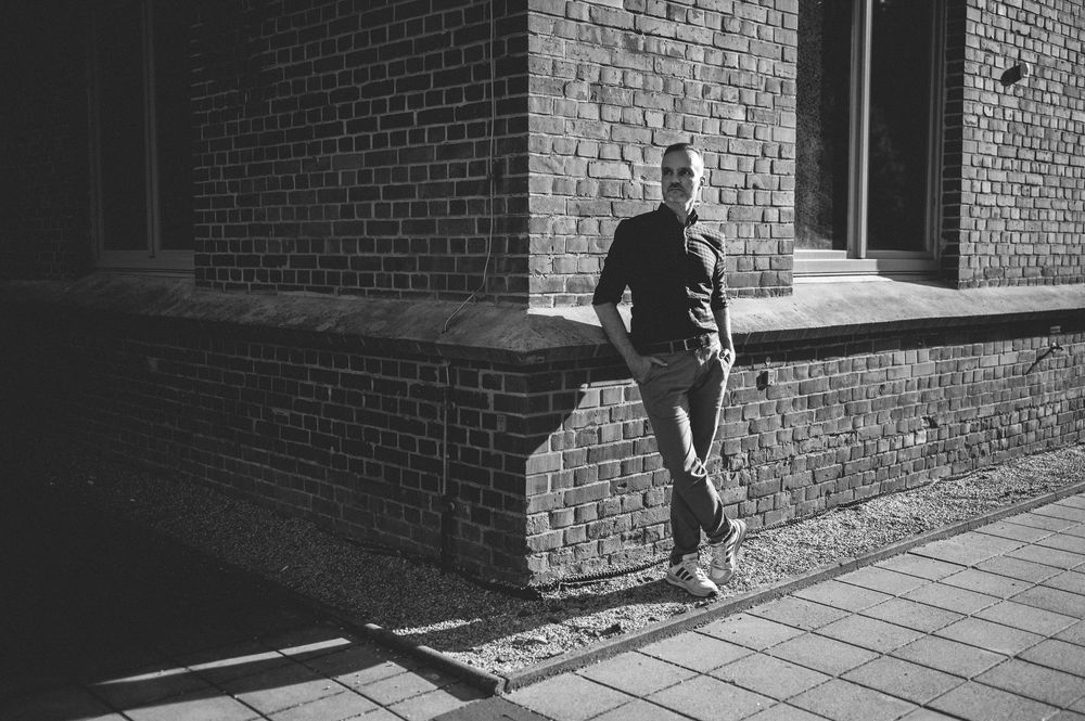 Person in dark clothing walking along a brick building exterior during daytime with harsh shadows.