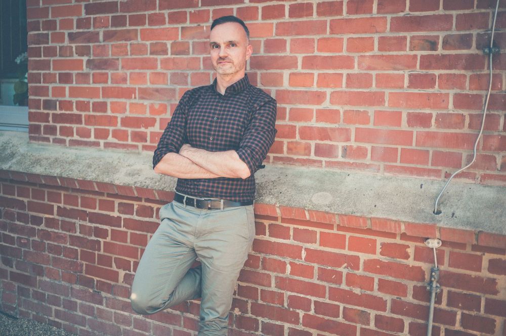Casual figure in plaid shirt and khakis leans against red brick wall on sunny day.