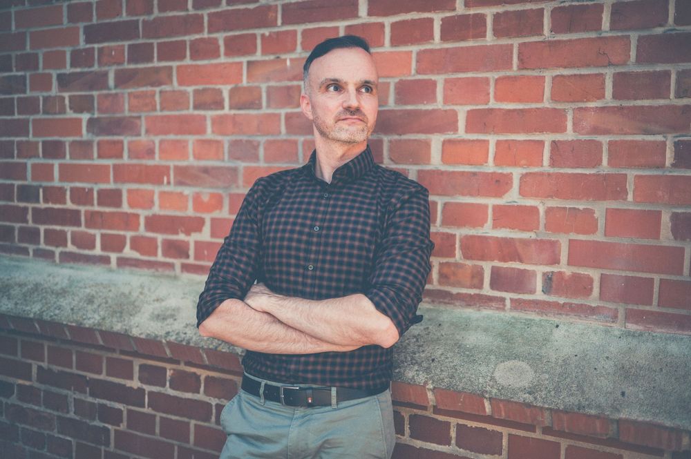 A person in a plaid shirt sits with crossed arms against a brick wall during a photoshoot session.