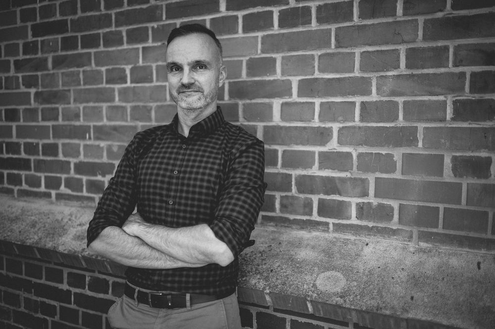 Professional headshot portrait against a brick wall in black and white.