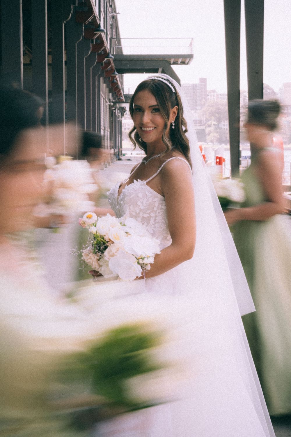 Elegant bride in white veil stands by wooden colonnade in soft light.