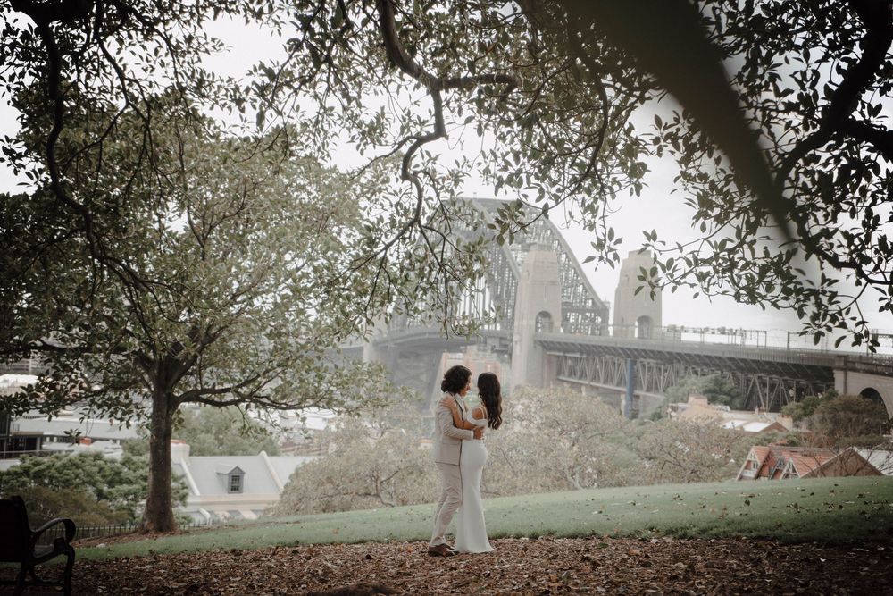 A couple embraces on a misty hillside with a stone cathedral visible in the background through overhanging trees.