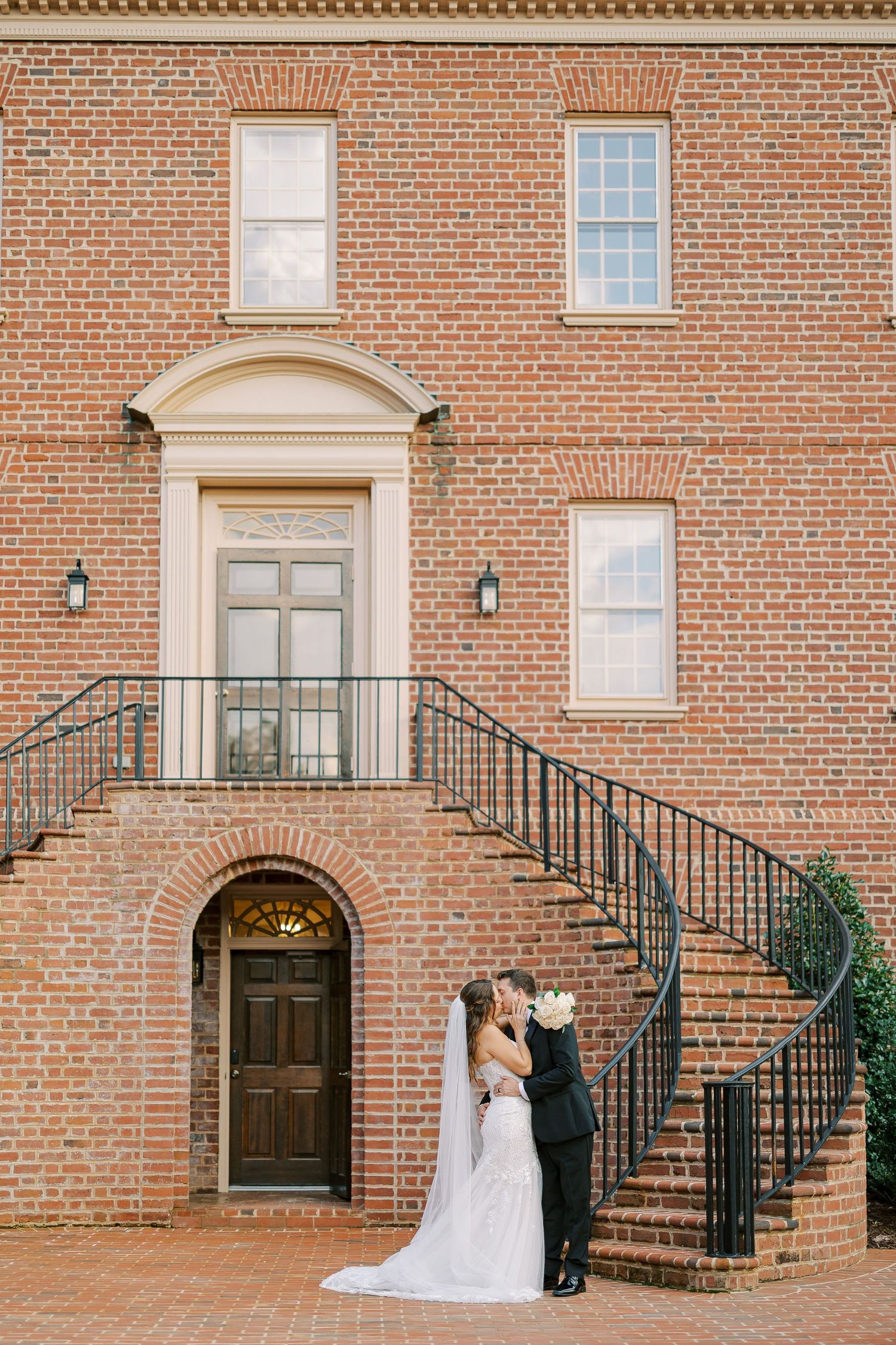 Bride and groom share a kiss on brick steps of elegant colonial building, bride holding white bouquet in flowing veil.