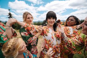 Bride in floral robe surrounded by bridesmaids in matching robes outdoors at a wedding celebration.