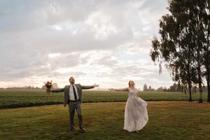Bride and groom joyfully tossing bouquet on green lawn at sunset during outdoor farm wedding celebration.