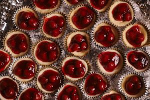Mini cherry cheesecake bites in foil cupcake liners topped with glossy red cherry pie filling on a baking sheet.