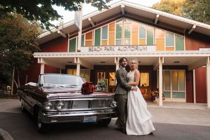 Bride and groom posing beside a vintage maroon car in front of Beach Park Auditorium on their wedding day.