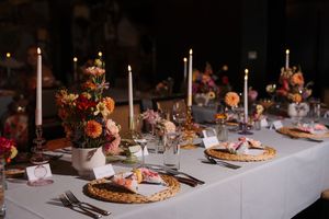 Elegant wedding reception table with candles, wicker chargers, and colorful floral centerpieces on a white linen tablecloth.